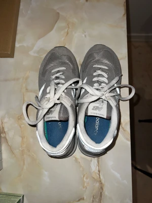 Grå New Balance 574 sneakers, stl 35 - Säljer ett par klassiska New Balance 574 sneakers i grått med vita detaljer. Skorna har snörning, rund tå och platt sula. Ovandelen är i mocka och mesh för en sportig vibe och skön komfort. Perfekta för dig som gillar stilrena och tidlösa sneakers.