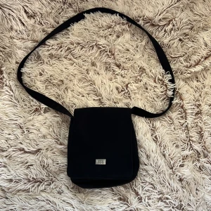 Svart axelväska från Zera Zera - Snygg svart axelväska från Zera Zera med stilren rektangulär form och lång axelrem. Väskan har ett lock med metallplatta där märket står, samt ett rymligt huvudfack och en ytterficka på baksidan. Perfekt för att bära med sig det viktigaste. Den har 4 fak.