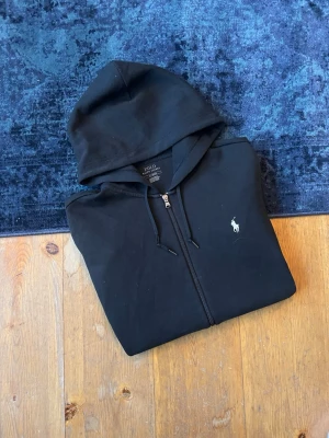 Svart Ralph Lauren Hoodie  - Hej, Säljer en svart Ralph Lauren zip hoodie med vit pony. Se skick enligt bilder, det finns tre riktigt små hål som endast gått igenom första lagret av hoodien. Den kommer i storlek M. Hör av er vid frågor!