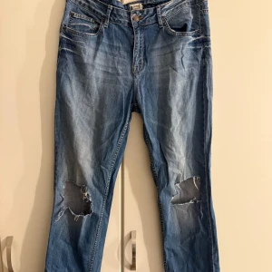 Blå Gina Tricot jeans med hål - Säljer ett par blå jeans från Gina Tricot, modell Perfect Jeans. De har slitna detaljer och stora hål vid knäna för en cool och avslappnad look. Klassisk femficksdesign och knappstängning framtill. Perfekt för dig som gillar streetstyle.