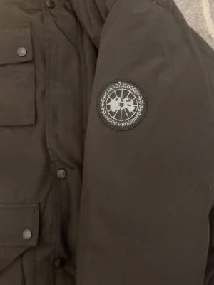 Canada goose jacka - Svart dunjacka från Canada Goose med klassisk patch på ärmen. Jackan har fyra stora fickor med knappar framtill och hög krage. Insidan är quiltad och jackan är tillverkad i polyester med dunfyllning. Perfekt för kalla vinterdagar.