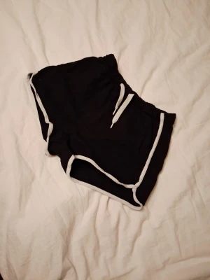 Svarta shorts med vit kant från H&M - Svarta shorts från H&M med vit kant och snörning i midjan. De har en loose passform och är gjorda i mjuk bomull, perfekta för chill dagar eller träning. Klassisk sportig look med elastisk midja och kontrastdetaljer.