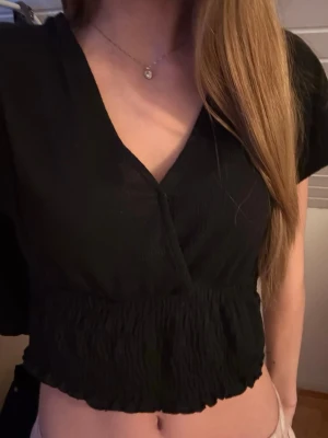 Svart v-ringad topp - En svart topp med omlott-design och v-ringad hals. Toppen har korta ärmar och ett elastiskt midjeparti med lätt volangkant. Skriv för bilder/frågor eller intresse!🩷