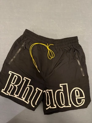Svarta Rhude shorts med gul snörning - Säljer ett par svarta shorts från Rhude i storlek M. De har stor vit logga framtill, gul snörning i midjan och dragkedjeförsedda fickor. Tillverkade i lätt syntetmaterial med meshfoder för extra komfort. Perfekta för en streetwear-look.