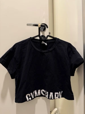 Svart Gymshark cropped t-shirt M - Svart cropped t-shirt från Gymshark i storlek M. Tillverkad i mjuk bomull med 5% elastan för extra stretch. T-shirten har rund hals och tryckt Gymshark-logga i vitt längs nederkanten. Perfekt för gymmet eller chill dagar.