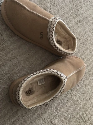 Beige UGG korta - Mysiga slip-in skor från UGG i beige mocka med fluffigt pälsfoder och dekorativt mönster runt öppningen. 