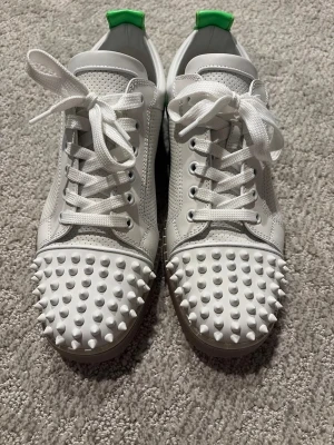 Christian Louboutin "ovanliga" - Säljer ett par vita sneakers från Christian Louboutin med gröna detaljer på hälen och röd sula. Skorna har snörning, perforerat skinn och coola vita nitar på tån. Märkets logga syns på insidan och hälen. Perfekt för dig som gillar statement pieces.