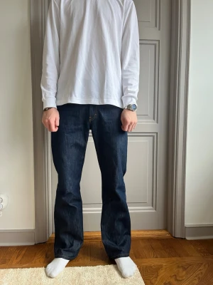 Jeanerica - Säljer ett par klassiska mörkblå Levis 501 jeans med rak passform. Jeansen har en tidlös look med fem fickor och gylf med knappar. Perfekta för dig som gillar en enkel och clean stil. Materialet är robust jeans i bomull.