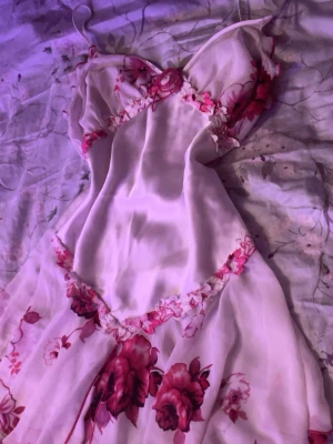 Blommig kortklänning med spetsdetaljer - Supersöt kortklänning i ljusrosa med blommigt mönster i rött och rosa. Klänningen har tunna axelband, spetsdetaljer och volangkant nertill. Materialet känns lätt och flowy, perfekt för varma dagar. Sitter snyggt med v-ringad urringning och markerad midja. Kan även användas som nattklänning 🌸 Från Palma🥰