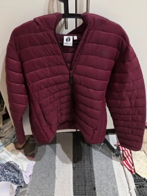 Vinröd pufferjacka från Hang Ten XL - Säljer en vinröd pufferjacka från Hang Ten i storlek XL. Jackan har quiltad design, dragkedja framtill och diskret logga på bröstet. Perfekt för kyliga dagar och riktigt skön att bära. Materialet är lätt och känns smidigt.