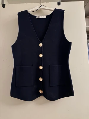 Mörkblå stickad väst från Zara - Snygg mörkblå stickad väst från Zara med v-ringning och stora guldfärgade knappar framtill samt  två  fickor. 