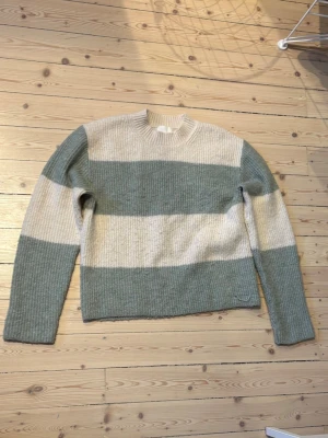 Randig stickad tröja i grönt och creme  - Säljer en mysig, oversized, stickad tröja med breda ränder i grönt och beige. Tröjan har rund hals och långa ärmar, perfekt för kyliga dagar. Den är ribbstickad och har en relaxed passform som gör den skön att bära.