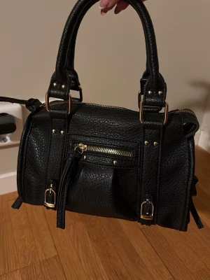 Svart handväska med gulddetaljer från Spanien  - Snygg svart handväska i skinnimitation med guldfärgade detaljer och dragkedja framtill. Väskan har dubbla handtag, justerbar axelrem och dekorativa spännen. Perfekt storlek för vardagens essentials och ger en cool vibe till din outfit.