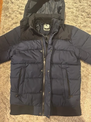 Mörkblå pufferjacka från McKinley - Snygg mörkblå pufferjacka från McKinley med svarta detaljer och huva. Jackan har dragkedja framtill, två sidofickor med dragkedja och ribbade muddar. Perfekt för kalla dagar och riktigt skön att bära.