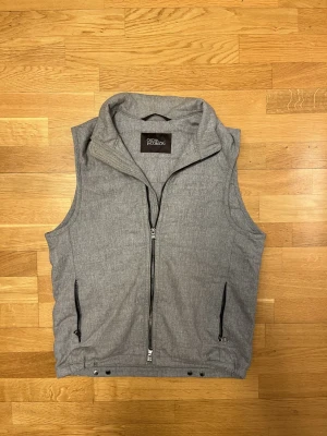 Oscar Jacobson flanellväst - Snygg grå flanellväst från Oscar Jacobson med hög krage och två dragkedjefickor framtill. Västen har en stilren look med hel dragkedja och knappar nertill. Perfekt att slänga över en hoodie eller tröja för extra värme. Storleken är 48/M 