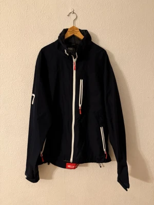 Vindjacka från Helly Hansen - Säljer en marinblå vindjacka från Helly Hansen med vita detaljer och dragkedjor. Jackan har hög krage, flera fickor med dragkedja och tryckta loggor på ärmen och kragen. Perfekt för blåsiga dagar och snygg till jeans eller träningskläder.