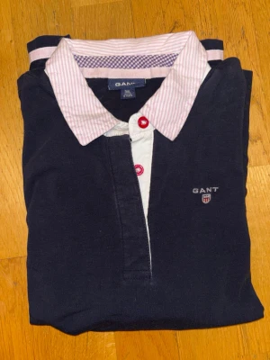 Mörkblå långärmad skjorta från GANT - Snygg mörkblå långärmad skjorta från GANT med vit och rosa randig krage och röda knappar. Skjortan har broderad logga på bröstet och är gjord i mjuk bomull. Perfekt för dig som gillar klassisk stil med en twist.