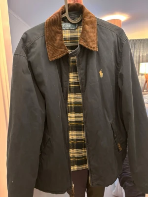 Mörkblå  vindjacka från Polo Ralph Lauren - Klassisk mörkblå vindjacka från Polo Ralph Lauren med brun manchesterkrage och gul broderad logga på bröstet. Insidan har rutigt foder i svart, gult och beige. Jackan har dragkedja framtill och två fickor. Typ aldrig använd. 