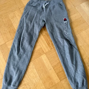 Grå mjukisbyxor från Champion ungdom - Grå mjukisbyxor från Champion med broderad logga längs benet och vit snörning i midjan. Tillverkade i mjukt bomullsmaterial med resår i benslut och midja för skön passform. Perfekta för chill eller träning.