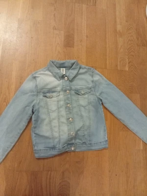 Ljusblå jeansjacka från &Denim - Snygg ljusblå jeansjacka från &Denim i klassisk modell med krage, långa ärmar och silverfärgade knappar framtill. Jackan har två bröstfickor med lock och knappar, samt raka sömmar som ger en clean look. Perfekt för dig som gillar street och casual vibes.