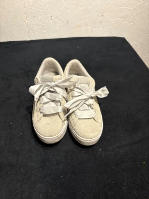 Beige sneakers från Puma med satinband - Snygga beige sneakers från Puma med breda satinband som snörning. Skorna har en rund tå och platt sula, tillverkade i mocka med klassisk Puma-logga på sidan. Perfekta för dig som gillar en clean och stilren look med en twist.