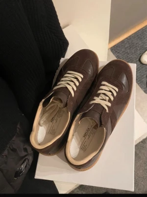 Maison Margiela Replica sneakers brun - Maison Margiela Replica sneakers i mörkbrunt skinn och mocka med beige snörning och gummisula. Klassisk rund tå och platt sula, med diskreta detaljer och stilren design. Perfekt för dig som gillar minimalistisk och tidlös stil.
