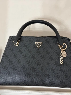 Guess väska - Säljer min Guess väska i färgen Coal (inte helt svart som det står). Sparsamt använd och i mycket fint skick utan slitage. Inget band medföljer🥰