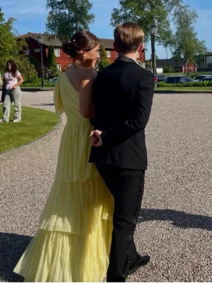 gul långklänning med lager på lager - Säljer en drömmig gul långklänning med lager på lager av volanger och oneshoulder-design. Unik då den är omsydd hos en skräddare då den från början har en slits, har lämnat in den på kemtvätt, jag på bilden är 172 och bärde klack på 4cm, den är verkligen drömmig🤩