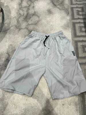 Grå loose shorts med snörning - Säljer ett par gråa shorts från Represent med loose passform och elastisk midja. De har svart snörning framtill och är tillverkade i ett lätt syntetmaterial som passar perfekt för varma dagar. Enkla och stilrena med en diskret logotyp på sidan.