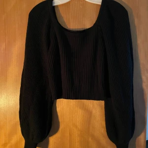 Svart croppad stickad topp - Säljer en svart croppad stickad topp med bred ribbad struktur och långa puffiga ärmar. Toppen har en snygg fyrkantig ringning som ger en cool vibe och passar perfekt till höga jeans eller kjol. Superenkel att styla för en trendig look.