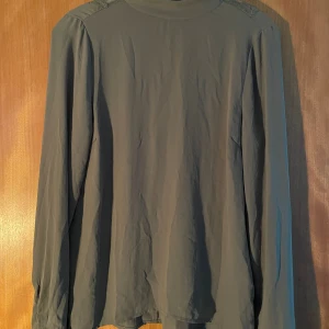 Grön blus med spetsdetaljer - Snygg grön blus med lång ärm och djup v-ringning baktill dekorerad med spets. Blusen har en lös passform och är perfekt för dig som gillar stilrena plagg med en twist. Materialet känns lätt och luftigt.