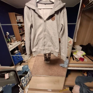 Grå hoodie med huva - Mysig grå hoodie i storlek S. Tröjan har huva, dragkedja framtill och två stora fickor. Insidan är fodrad med mjuk fleece och det finns snygga bruna detaljer på bröstet och ärmen. Perfekt för chill dagar och när du vill vara extra bekväm.