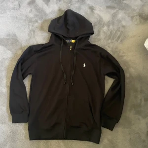 Svart hoodie från Polo Ralph Lauren - Svart hoodie från Polo Ralph Lauren med dragkedja och huva. Klassisk design med vit broderad logga på bröstet, känguruficka och snörning i huvan. Tillverkad i mjuk bomull, perfekt för chill dagar eller när du vill ha en clean look.