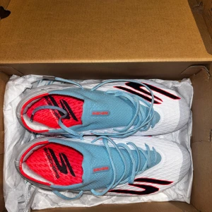 Skechers vita och blå fotbollsskor - Säljer ett par Skechers fotbollsskor med snygg design i vitt, ljusblått och rött. Skorna har snörning, rund tå och platt sula med röda dobbar. Ovandelen är tillverkad i syntetmaterial och har svarta detaljer på sidan samt röd insida med Skechers-logga.