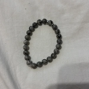 Gråmelerat pärlarmband med stenmönster - Snyggt armband med runda pärlor i gråmelerad stenlook och svarta marmoreringar. Elastiskt band gör det enkelt att ta på och av. Perfekt accessoar för dig som gillar stilrena och unika detaljer.