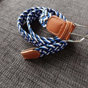 Randigt flätat bälte med skinn-detaljer - Säljer ett snyggt flätat bälte med randigt mönster i blått, svart och vitt. Bältet har bruna skinn-detaljer vid spännet och ändarna, samt ett silvrigt metallspänne. Perfekt för att ge outfiten lite extra edge och färg.