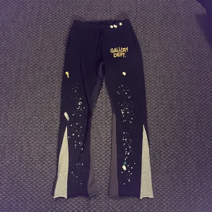 Svarta Gallery Dept. mjukisbyxor med färgstänk - Unika svarta mjukisbyxor från Gallery Dept. med grå paneler längs benen och coola färgstänk i vitt, gult och blått. Byxorna har resår i midjan, snörning och tryckt logga framtill. Tillverkade i 100% bomull för maximal komfort.