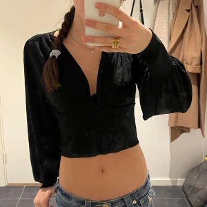 Svart croppad blus med v-ringning - Säljer en svart croppad blus med djup v-ringning och långa puffärmar. Toppen har knappar framtill och ett diskret blommigt mönster i tyget som ger en snygg textur. Perfekt för dig som gillar stilrena och trendiga plagg med lite extra detaljer.