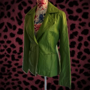 Grön skinnjacka från Vero Moda - Snygg grön jacka i skinn från Vero Moda med klassisk krage, knappstängning och två stora fickor framtill. Jackan har lång ärm och en stilren, figurnära passform som ger en cool och unik look. Perfekt för dig som vill sticka ut med färg!