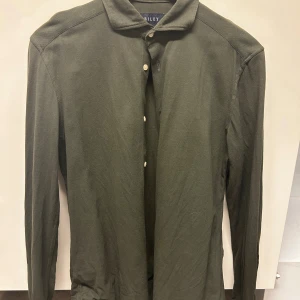 Olivgrön henley-skjorta från Riley - Snygg olivgrön henley-skjorta från Riley i slim fit. Skjortan har lång ärm, klassisk krage och knappar framtill. Perfekt för dig som gillar stilrena och enkla plagg med en modern passform.