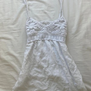 Vit virkad linnetopp med spetsdetaljer - Söt vit linnetopp med virkade spetsdetaljer upptill och tunna axelband. Toppen har en luftig passform och är perfekt för varma dagar. Virkningen upptill har blommönster och ger en bohemisk känsla.