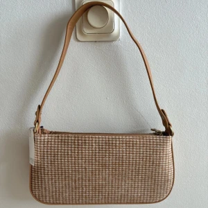 Beige rutig axelväska från Lindex - Snygg axelväska från Lindex i beige och vitt rutigt mönster. Väskan har en mjuk, rektangulär form med justerbart axelband i beige syntetmaterial och dragkedja upptill. Perfekt storlek för det viktigaste och med innerfack för småsaker. Helt ny med prislapp! Nypris: 255kr