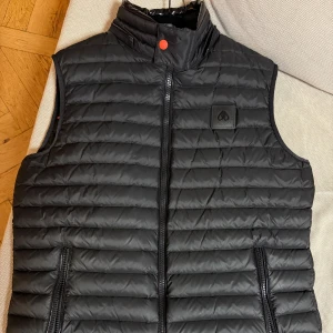 Svart dunväst från Moose Knuckles - Snygg svart dunväst med quiltad design och hög krage. Västen har dragkedja framtill, två sidofickor med dragkedja och en diskret logotyp på bröstet. Perfekt för lager-på-lager och chill stil. Luvan saknas !