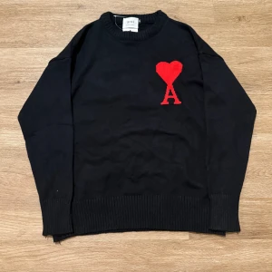 Svart stickad tröja från Ami - Svart stickad tröja från Ami med rund hals och ribbade muddar. På bröstet finns ett rött hjärta och ett stort A, vilket ger en unik och snygg detalj. Perfekt för dig som gillar stilrena plagg med en twist.