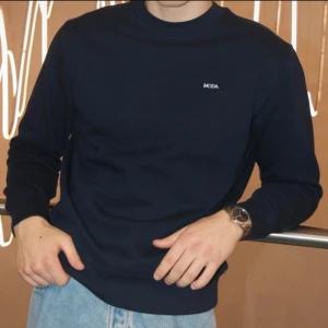 Mörkblå sweatshirt  - En stilren mörkblå sweatshirt från Stanley/Stella med liten vit MODA-logga på bröstet. Tröjan har rund hals, långa ärmar och är tillverkad i mjuk bomull. Perfekt för en clean och avslappnad look.