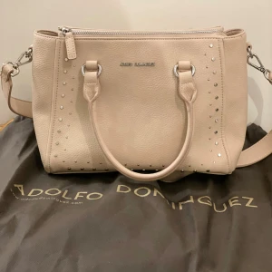  Väska från Adolfo Dominguez - Snygg beige väska från Adolfo Dominguez i  skinn med silverfärgade nitar längs sidorna. Väskan har både handtag och axelrem, samt dragkedja upptill. Aldrig använd.