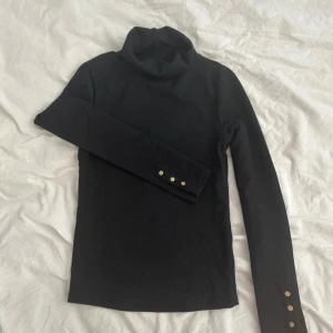 Svart polotröja från ASOS DESIGN - Svart polotröja från ASOS DESIGN med lång ärm och snygga guldfärgade knappar vid ärmsluten. Perfekt för dig som gillar stilrena och klassiska plagg med en liten twist. Polokragen ger extra cozy vibe och passar till många olika looks.