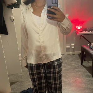 Vit satinliknande skjorta med knappar - Snygg vit skjorta i satinliknande material med klassisk krage och långa ärmar. Skjortan har en rak passform och stängs med knappar framtill. Perfekt för dig som gillar en clean och stilren look.