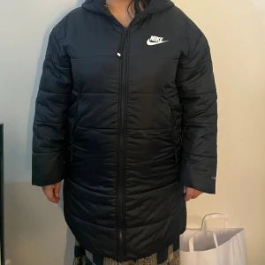 Svart lång pufferjacka från Nike - Svart lång pufferjacka från Nike. Jackan har huva, dragkedja framtill och två sidofickor med dragkedja. I storlek L