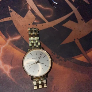 Guldfärgad klocka från Michael Kors - Stilren klocka från Michael Kors med rund urtavla i silver och guldfärgat länkarmband i rostfritt stål. Urtavlan är minimalistisk med tunna svarta markeringar och tre visare. Perfekt accessoar för dig som gillar klassisk design med lyxig känsla. Behövs nytt batteri!